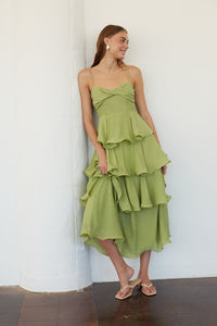 A woman wearing a light olive green strapless ruffled midi dress with a twisted sweetheart bodice, leaning against a white wall.