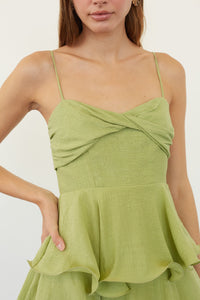 A woman wearing a light olive green spaghetti-strap top with a twisted criss-cross bodice and a ruffled peplum hem, shown against a plain off-white background.