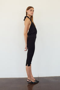 A woman stands in profile wearing a black sleeveless halter-neck top and matching black capri-length skirt or dress, paired with black pointed flats, against a plain white wall and dark floor.
