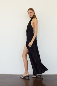 A woman models a black halter-neck maxi dress with a high thigh slit and draped side detail, paired with black thong sandals, standing against a plain white wall.