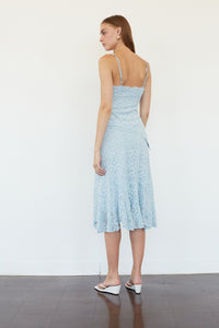 A woman stands with her back to the camera wearing a light blue lace midi dress with spaghetti straps and white wedge sandals against a plain white wall.