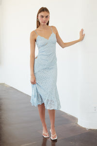A woman modeling a light blue lace spaghetti-strap midi dress in a minimalist white-walled room with dark wood floors.