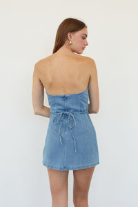 A woman shown from the back wearing a light blue denim strapless mini dress with a tie detail at the waist, set against a plain white studio background.