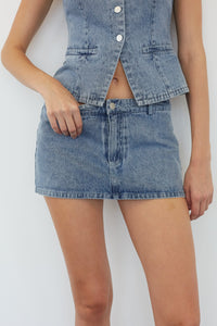 A model wearing a light blue denim set consisting of a cropped button-front vest and a matching denim mini skirt, shown from the chest down against a plain light background.