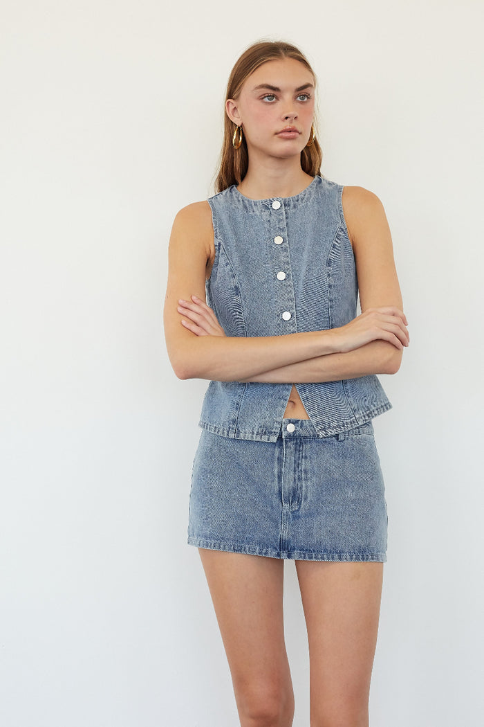 A model wearing a light blue denim sleeveless button-front top and matching denim mini skirt, standing against a plain white studio background.