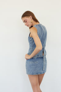 A woman wearing a sleeveless light blue denim mini dress, shown from a side-back angle against a plain white studio background.