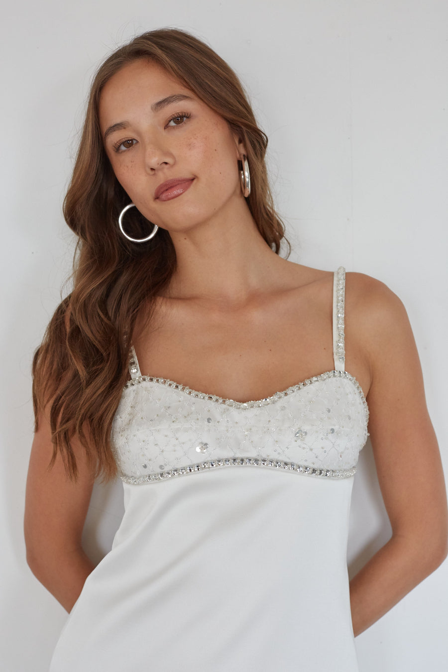 Woman wearing a white dress with thin straps and embellishments against a plain background