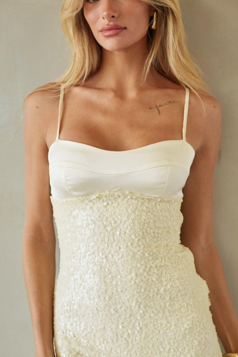 A woman wearing a cream-colored spaghetti-strap dress with a smooth satin-like bodice and a heavily textured, shimmering sequined or glittery waist and skirt, photographed against a neutral beige background.
