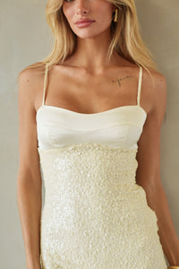 A woman wearing a cream-colored spaghetti-strap dress with a smooth satin-like bodice and a heavily textured, shimmering sequined or glittery waist and skirt, photographed against a neutral beige background.