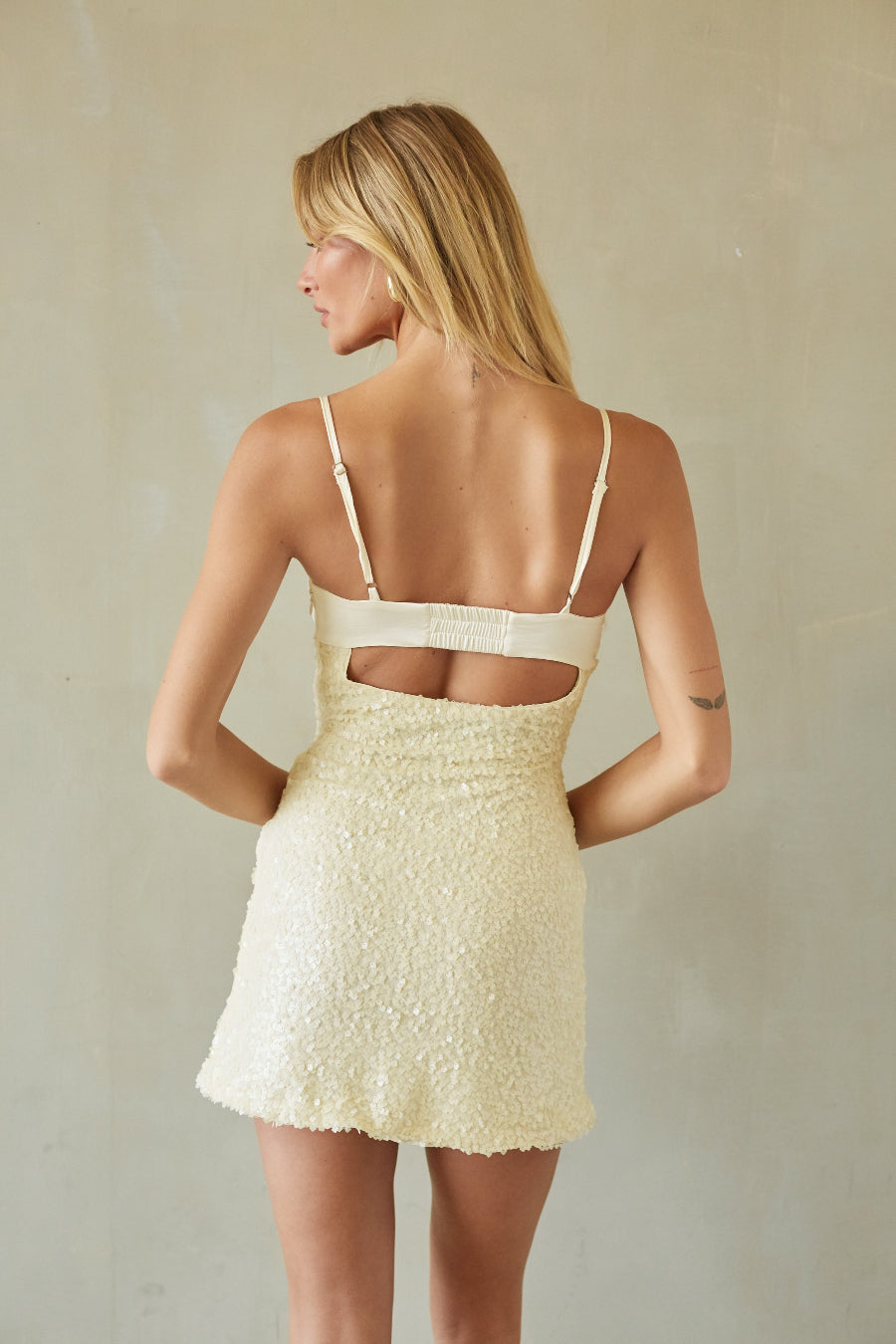 A woman shown from the back wearing a textured cream mini dress with thin straps and a cutout at the back, standing against a neutral beige wall.