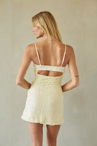 A woman shown from the back wearing a textured cream mini dress with thin straps and a cutout at the back, standing against a neutral beige wall.