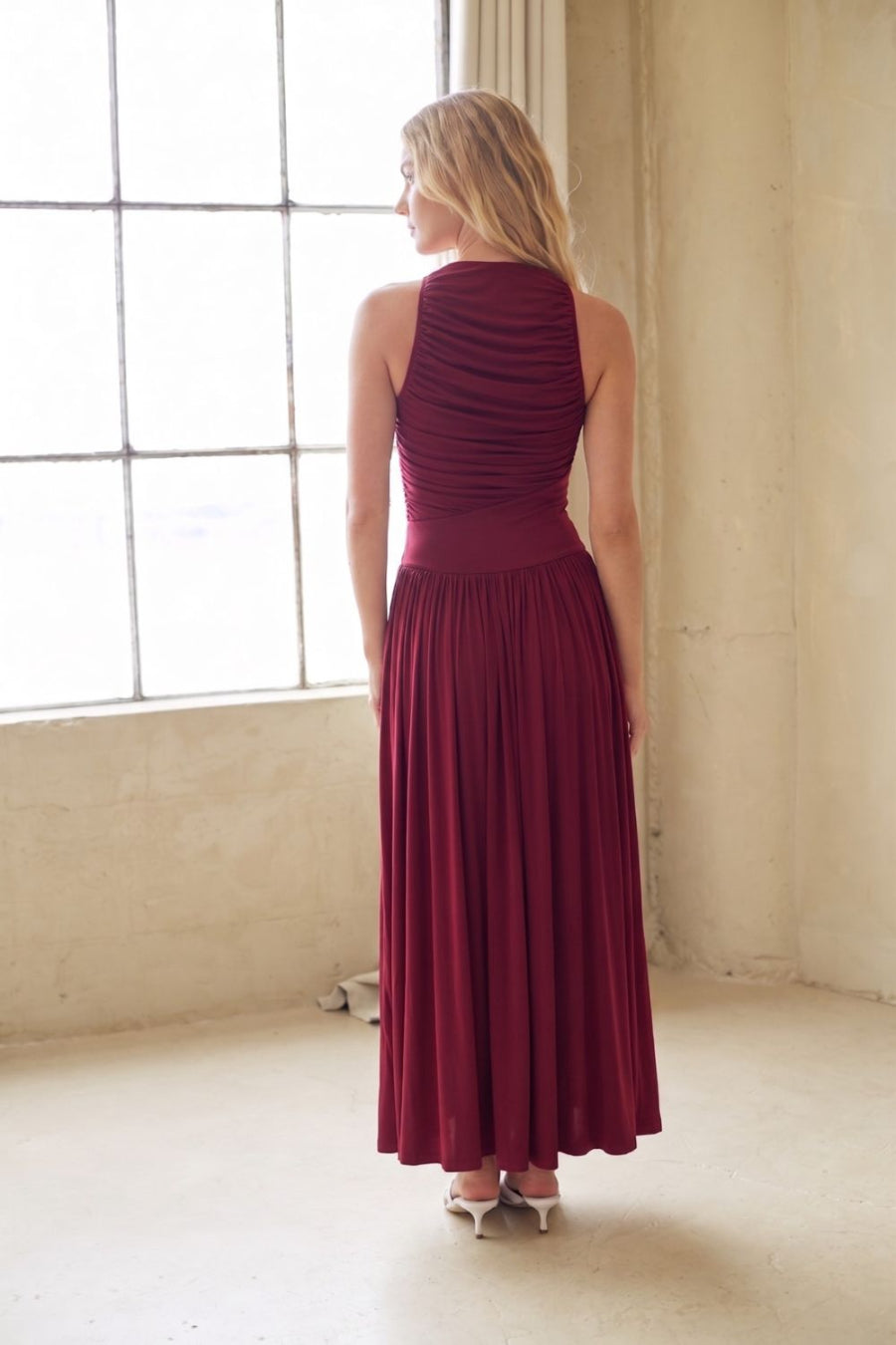 A woman stands with her back to the camera wearing a sleeveless, high-neck, floor-length pleated dress in deep burgundy, set in a bright, sunlit studio with a large window.