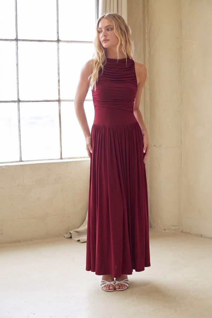 red-image | A woman modeling a sleeveless, high-neck, floor-length maxi dress in deep burgundy, standing in a bright, minimalist studio with a large window and concrete walls.