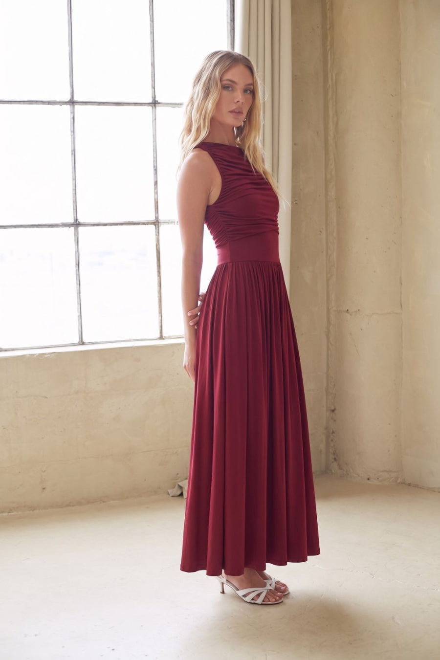 A woman modeling a sleeveless, high-neck, floor-length burgundy maxi dress in a bright, sunlit room with a large window and textured beige walls.