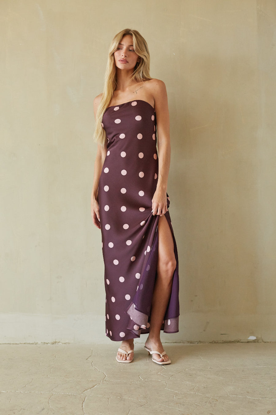 A woman modeling a strapless maxi dress with a purple base and white polka dots, standing against a textured beige plaster wall.