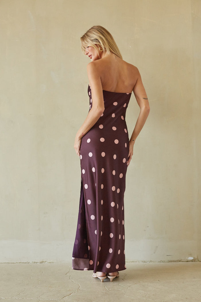 A woman wearing a strapless floor-length gown with large pink polka dots on a deep plum-purple background, photographed from the back against a textured beige wall.