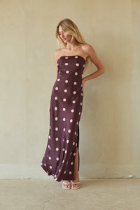 A woman modeling a strapless maxi dress with a dark plum purple base and light pink polka dots, standing against a textured beige plaster wall.