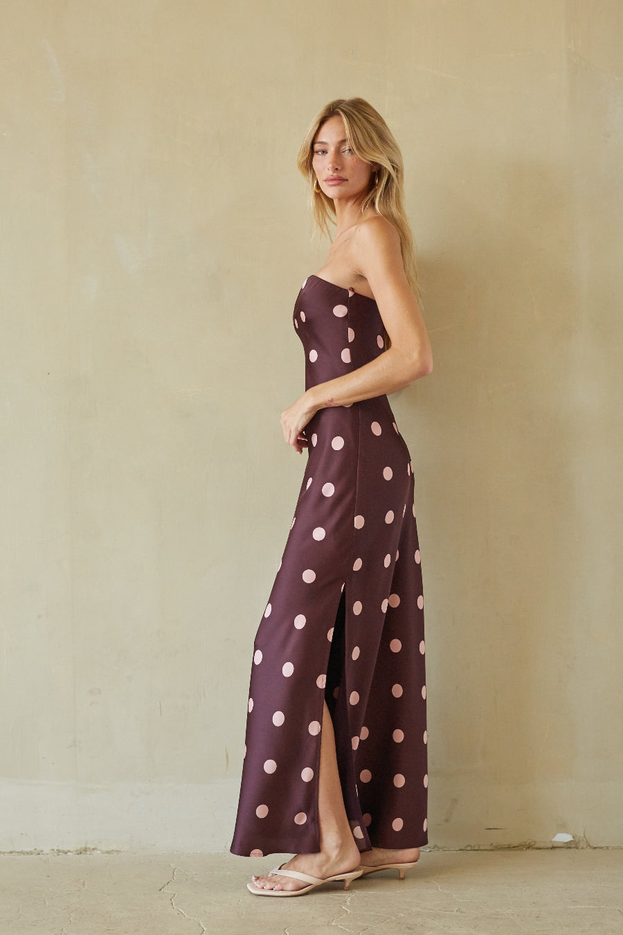 A woman modeling a strapless maxi dress with a dark plum base and large light pink polka dots, standing against a textured beige plaster wall.