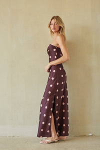 A woman modeling a strapless maxi dress with a dark plum base and large light pink polka dots, standing against a textured beige plaster wall.