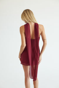 A woman viewed from behind wearing a sleeveless burgundy wrap dress with a long draped back panel and a high-low hem, set against a plain white studio background.