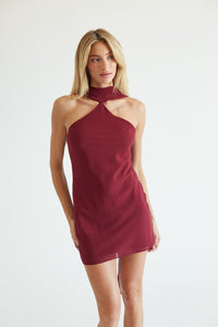 A woman modeling a sleeveless halter-neck mini dress in a deep burgundy color, set against a plain light gray studio background.