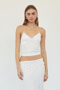 A woman modeling a white two-piece outfit consisting of a sleeveless crop top with thin straps and a matching long skirt with a polka dot pattern, standing against a plain white wall.