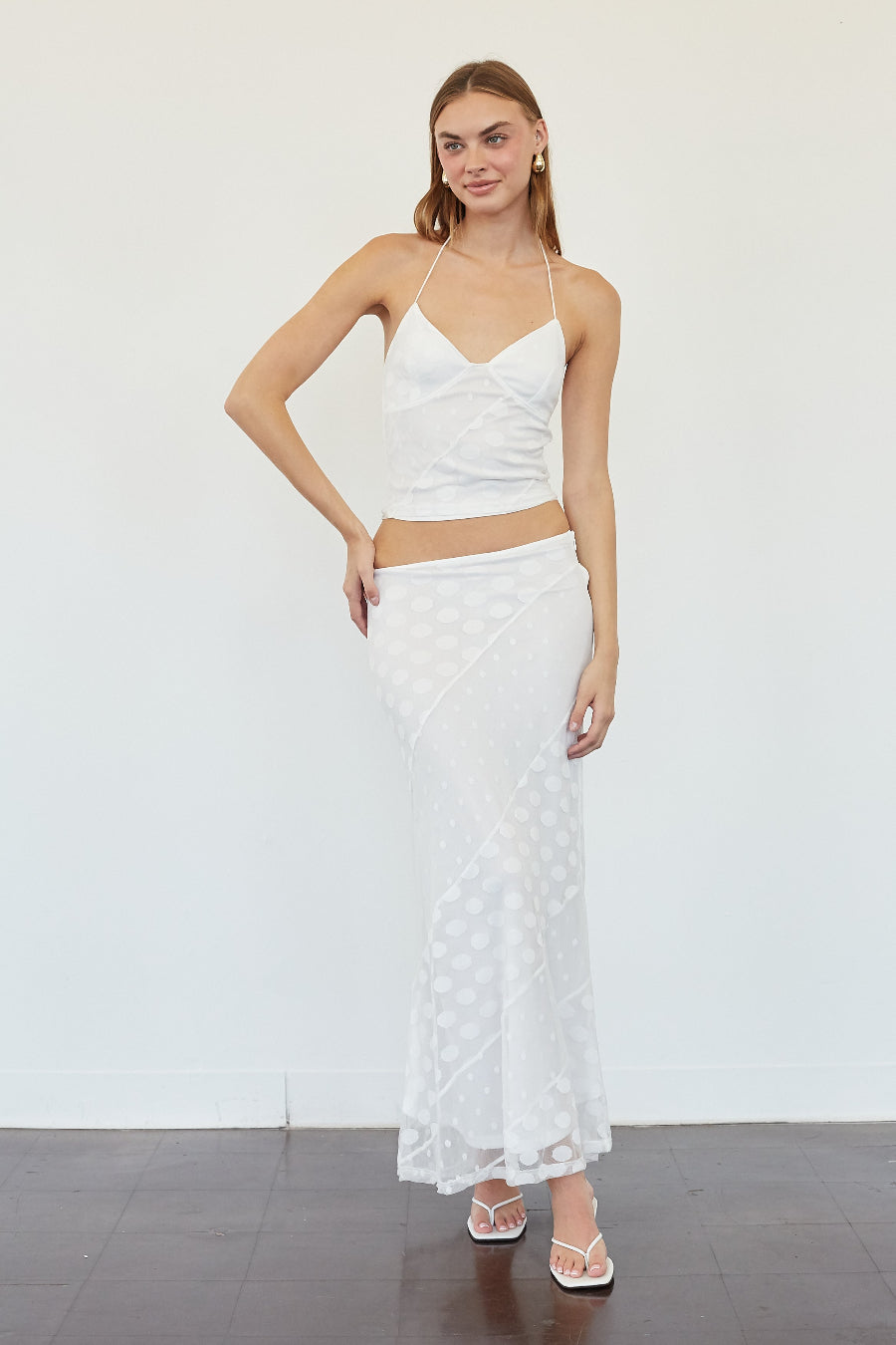 A woman modeling a white two-piece outfit consisting of a spaghetti-strap crop top and a matching textured midi skirt, standing against a plain white wall.