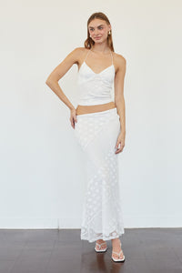 A woman modeling a white two-piece outfit consisting of a spaghetti-strap crop top and a matching textured midi skirt, standing against a plain white wall.