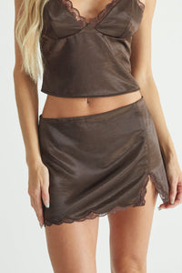 brown-image | A blonde model wearing a dark brown satin two-piece set consisting of a halter crop top with lace trim and a matching mini skirt, photographed against a light gray studio background.