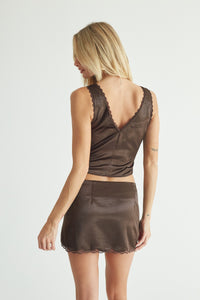 A woman shown from the back wearing a matching brown satin-like two-piece set with a scoop-neck crop top and a short skirt, both featuring scalloped edges, against a plain light gray studio background.