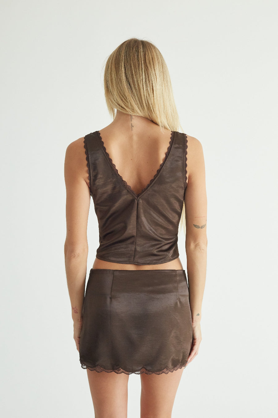 A woman shown from the back wearing a dark brown two-piece set consisting of a sleeveless crop top with a scalloped V-neckline and a matching mini skirt with a scalloped hem, against a plain light gray background.