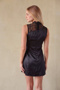 A woman wearing a black satin-like sleeveless dress with black lace shoulder panels, shown from the back against a plain beige wall.