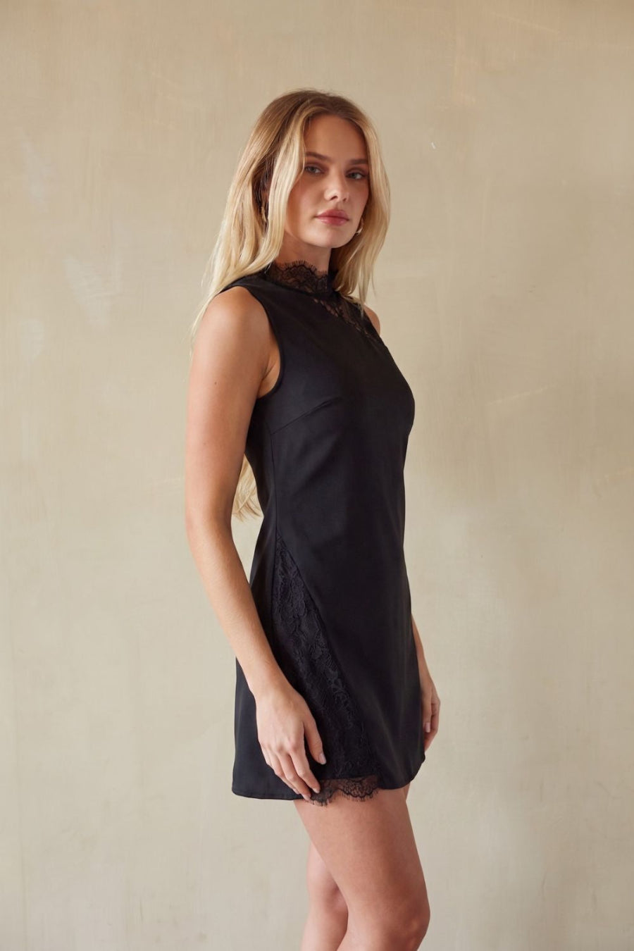 A blonde woman modeling a sleeveless black mini dress with lace trim at the neckline and hem, standing against a textured beige wall.