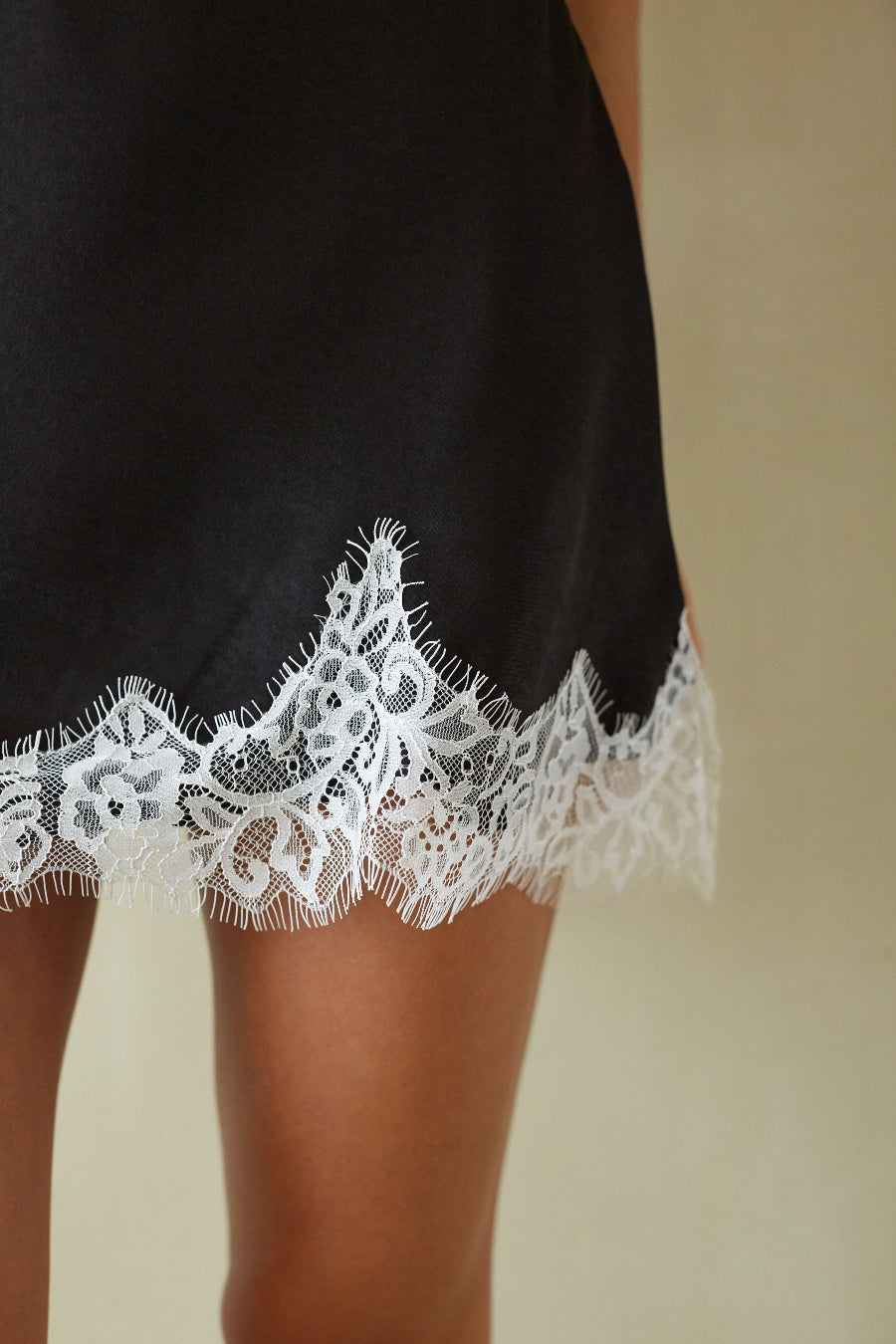 Close-up of a black slip dress or nightgown with a white floral lace hem, worn by a model against a neutral beige background.
