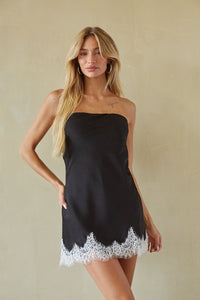 A blonde model wearing a black strapless tube dress with a white lace hem, standing against a textured beige wall.