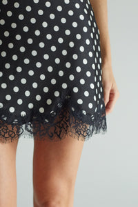Close-up of a black and white polka dot skirt with a black lace hem, worn by a model against a light gray background.