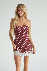 A blonde model wearing a strapless red and white polka dot dress with white lace trim, standing against a light gray studio background.