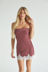 A blonde model wearing a strapless red and white polka dot dress with a white lace hem, standing against a light gray studio background.