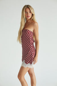 A blonde model wearing a strapless red dress with white polka dots and a white lace hem, standing against a light gray studio background.