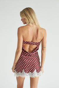 A blonde model wearing a strapless red dress with white polka dots and a white lace hem, open back, standing against a light gray studio background.