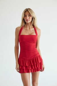 A blonde model wearing a bright red halter-neck mini dress with a square neckline, ruched bodice, and a tiered ruffled hem, set against a plain light gray studio background.