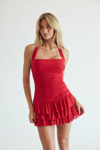 A blonde model wearing a bright red halter-neck mini dress with a square neckline, ruched bodice, and a tiered ruffled skirt, photographed against a plain light gray studio background.