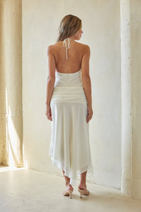 Woman wearing a white dress standing in a softly lit room with curtains.