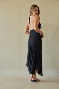 Woman wearing a black dress with a halter neck and flowing skirt against a beige wall.