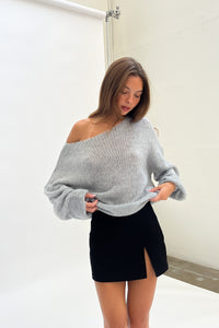 Woman wearing a gray off-shoulder sweater and black skirt against a white wall.