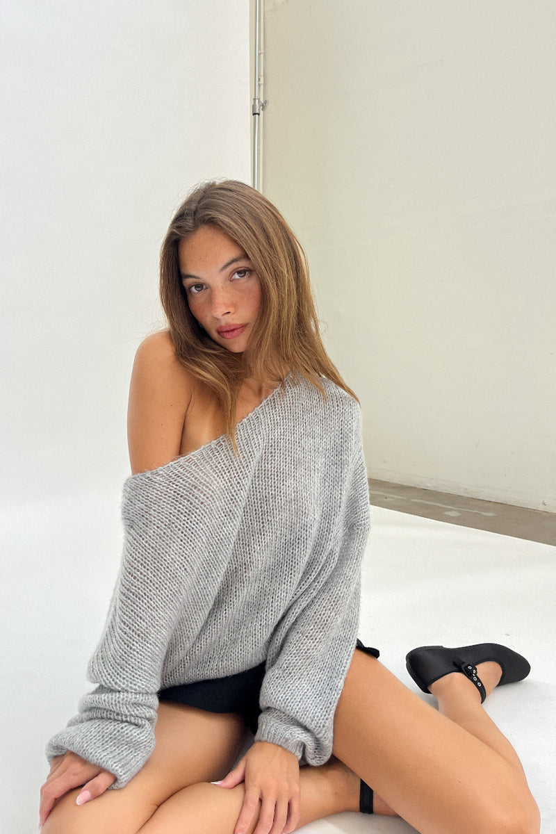 Woman wearing a gray off-shoulder sweater sitting on a white surface.