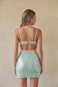 A woman shown from the back wearing a mint green sequined mini dress with thin straps and an open back, standing against a plain beige wall.
