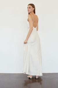 A woman modeling a floor-length white satin slip dress with thin straps and an open back, shown from the back in a minimalist studio setting.