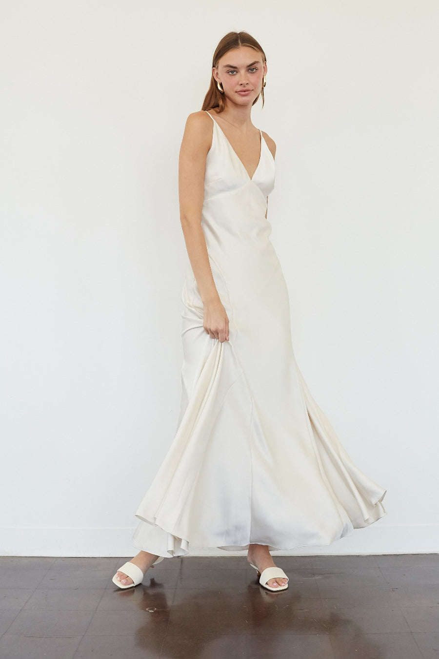 A woman modeling a sleeveless, off-white satin maxi dress with a deep V-neckline and flowing skirt, standing against a plain white wall.