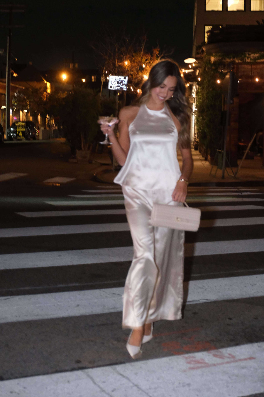 Woman in a white satin outfit crossing a street at night.